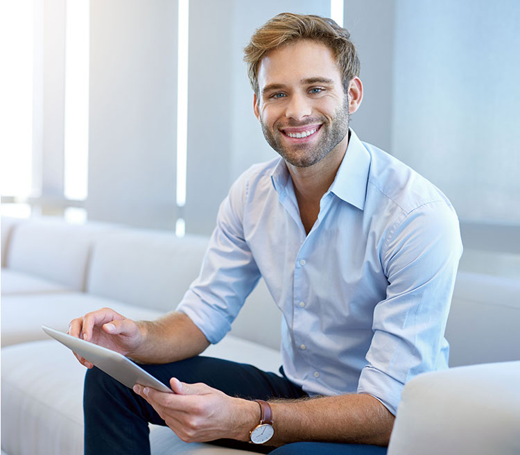 A smiling man holding a tablet sits casually in a well-lit room with modern furniture.